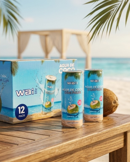 Presentación premium Wai Life de agua de coco con pulpa en playa tropical con caja de 12 pack, latas y cocos naturales sobre mesa de madera frente al mar.